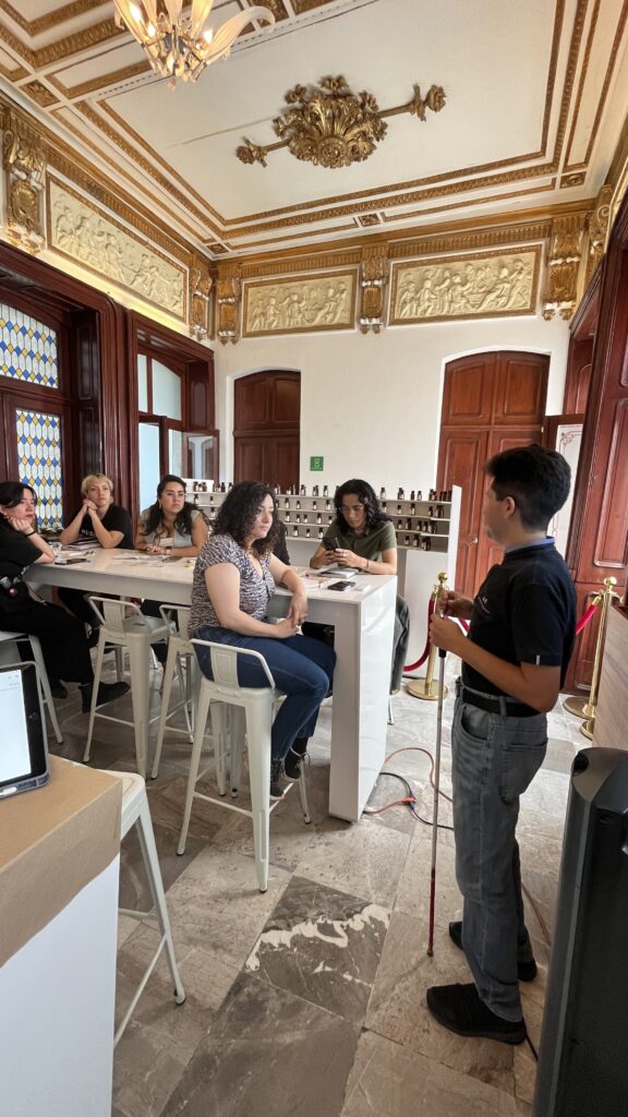 Foto de un hombre con discapacidad visual impartiendo una clase a un Equipo de guías del museo del perfume. Están en un salón de un edificio antiguo, con muchos detalles esculpidos en el techo.